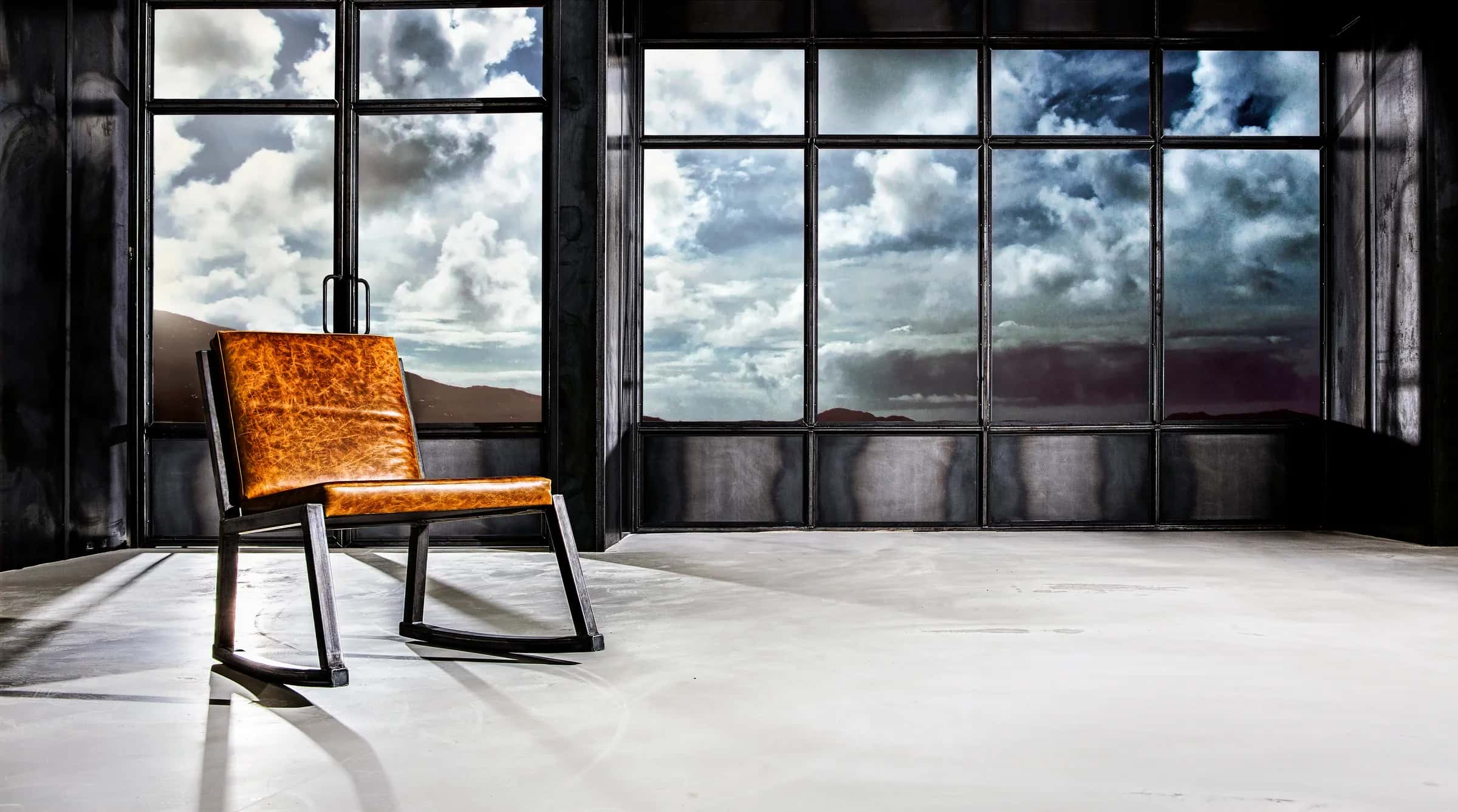 Cognac leather chair on a black steel frame in front of a tall steel window with clouds behind it